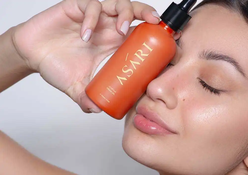 A close-up of a woman holding a bottle of Asari Beauty Argan Oil next to her face, showcasing radiant skin and a natural glow.